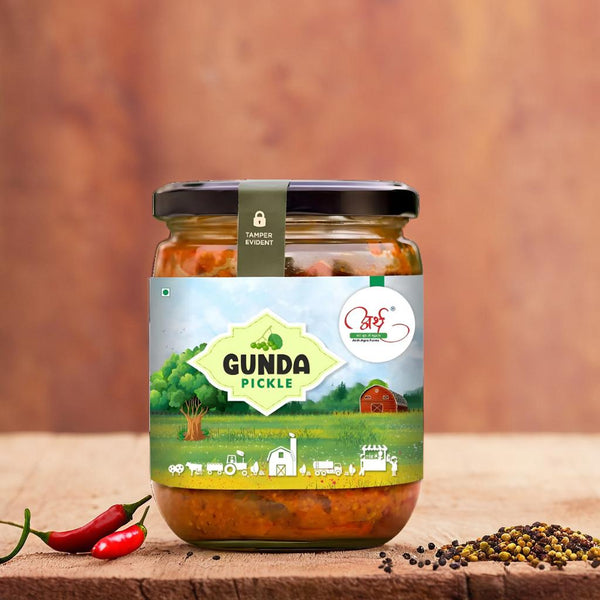 A jar of Authentic Gunda Pickle placed on a wooden surface, with a label depicting the product name and an illustration of a field with plants. Peppers and mustard seeds are visible in the foreground.