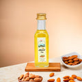 A clear glass bottle of wood-pressed almond oil with a gold cap, labeled with the product name and brand. The bottle is accompanied by almonds on a wooden board and a small bowl of almonds is visible in the background.